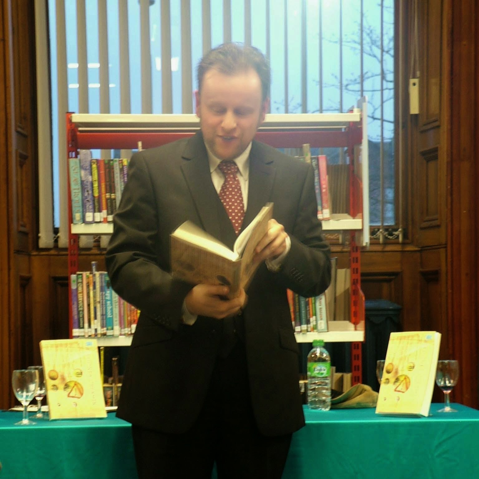 Me in a suit and tie reading from a book while in front of a table displaying said book