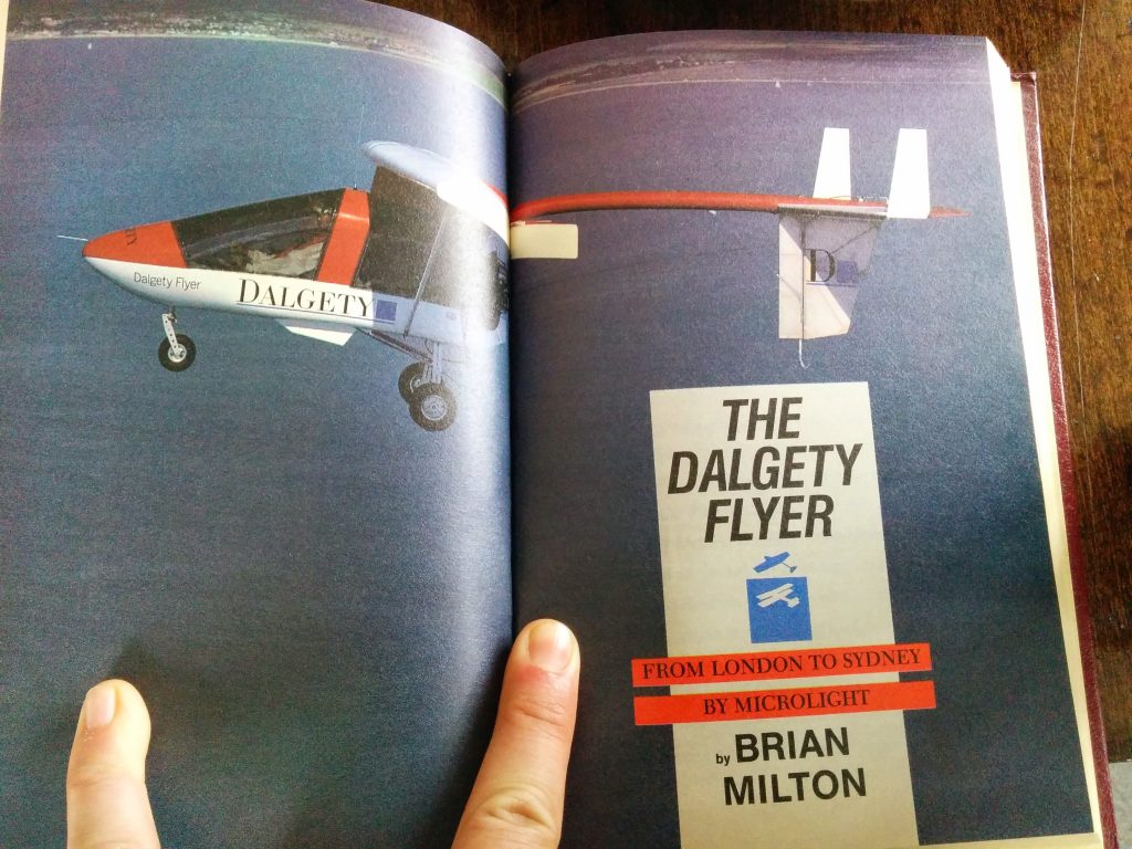 Two fingers hold open a book. Across two pages is the image of a microlite plane flying over water. On the righthand page is the title of a book "The Dalgety Flyer, from London to Sydney by microlite by Brian Milton"