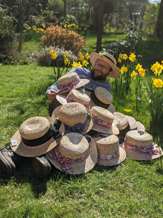 Myself, a bearded white man in glasses, lying on the grass in the sun. Behind me are bright yellow daffodils. I am wearing a straw boater hat and am covered in many other straw boater hats.