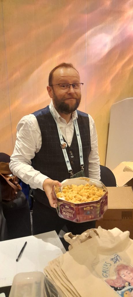 Me in a white shirt and blue waistcoat holding a tin full of tablet over a table with some Gallus tote bags on it