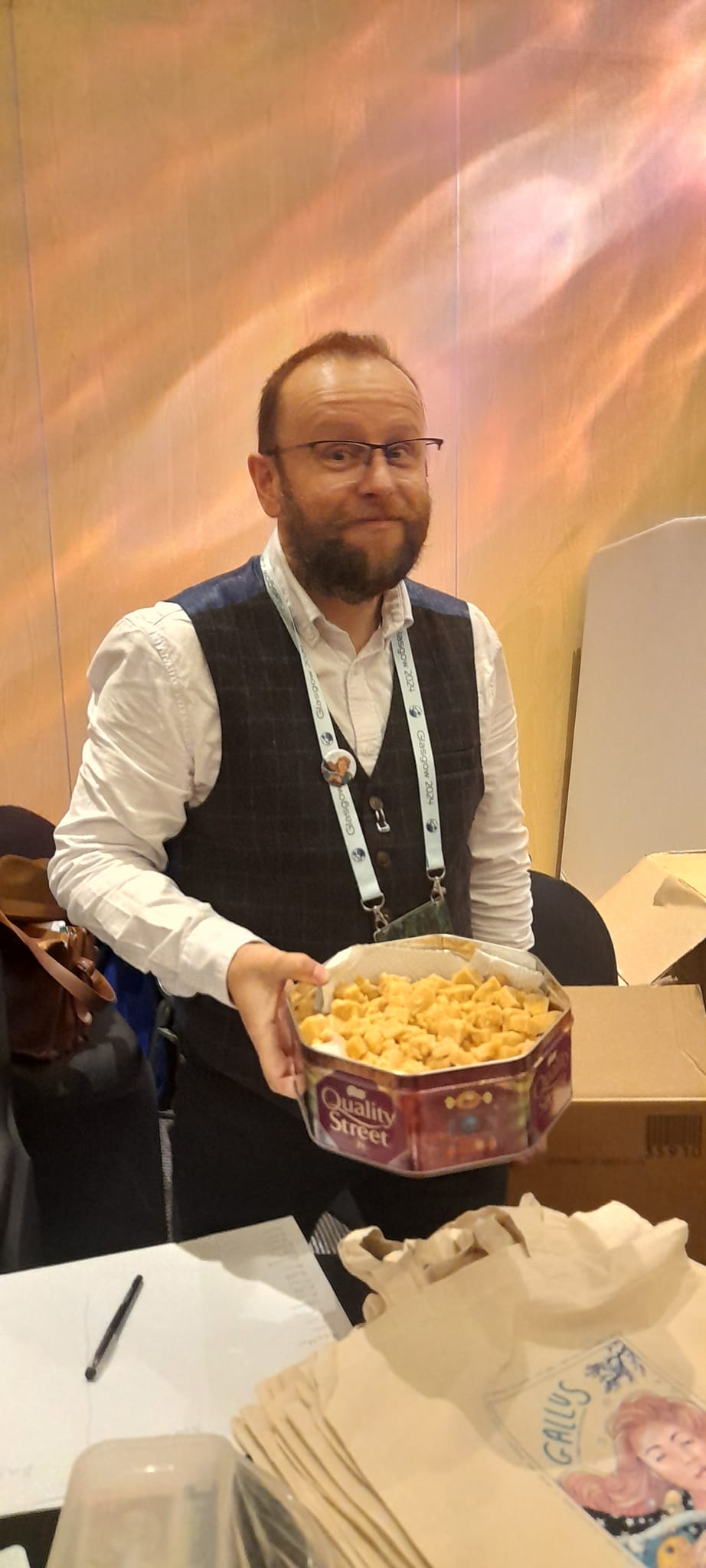 Me in a white shirt and blue waistcoat holding a tin full of tablet over a table with some Gallus tote bags on it 